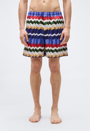 Άνδρας που φοράει πολύχρωμα σορτς με γεωμετρικό σχέδιο, όρθιος ξυπόλητος μπροστά σε απλό φόντο.