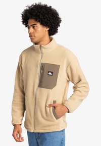 Beige fleecjacka med hög krage, dragkedja fram och ett brunt bröstfack. Har sidfickor och en mjuk, strukturerad yta.