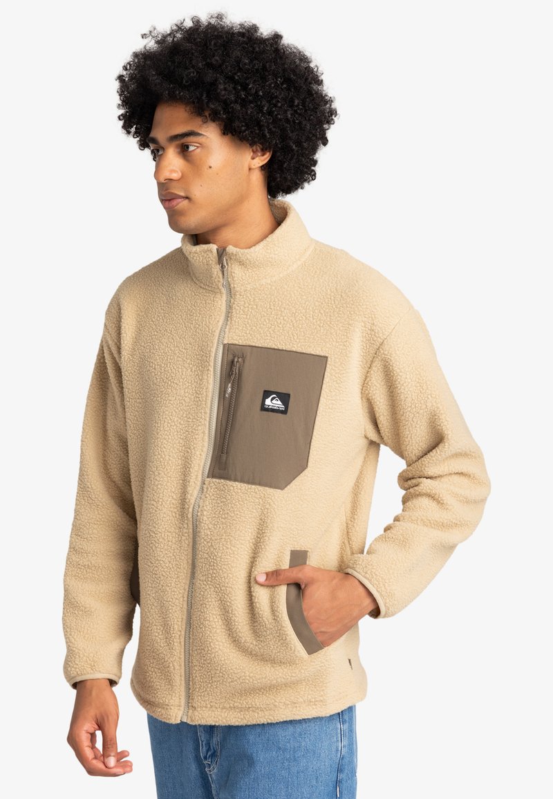 Beige fleecjacka med hög krage, dragkedja fram och ett brunt bröstfack. Har sidfickor och en mjuk, strukturerad yta.