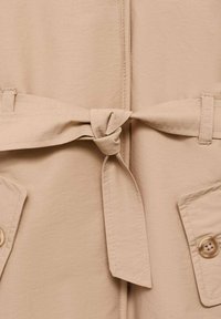 Ceinture en tissu beige nouée sur un vêtement avec deux poches boutonnées, mettant en valeur les détails de couture et de texture.