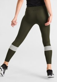 Olivově zelené legíny s vysokým pasem, zdobené texturovaným bílým vzorem na dolních nohavicích. Vyrobené z pružného, lehkého materiálu.