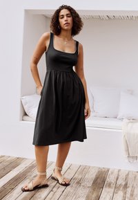 Fekete ujjatlan ruha, testhezálló felsőrésszel és ráncolt szoknyával. Négyzet alakú nyakkivágással és oldalzsebekkel rendelkezik. Bézs szandállal párosítva.