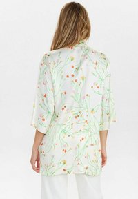 Femme aux cheveux longs portant un chemisier blanc à motifs floraux avec des tiges vertes et des fleurs orange, jaunes et roses, se tenant de dos.