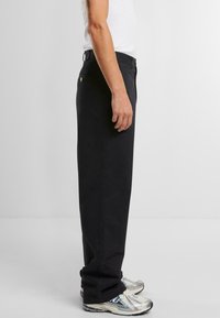 Black straight-leg pants made of cotton, featuring a back pocket with a small logo, worn with silver sneakers. Minimalist design and texture.