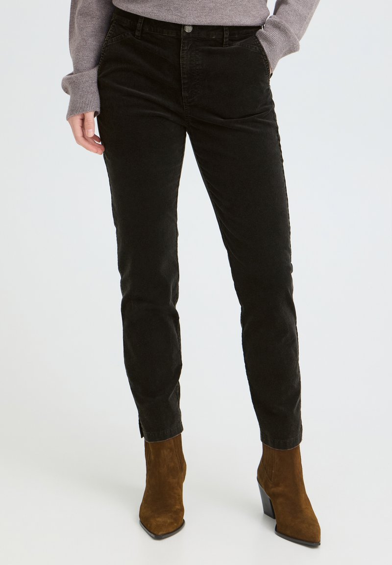 Femme portant un pantalon en velours côtelé noir slim, associé à des bottines en daim marron et un pull gris à manches longues.