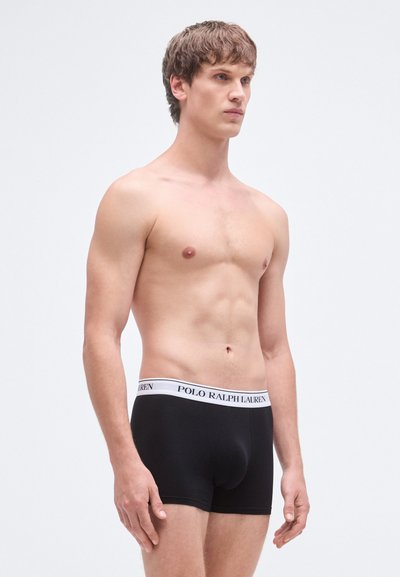 Cuecas boxer de algodão pretas com cintura elástica branca com o texto "POLO RALPH LAUREN" impresso. Modelo em posição neutra.