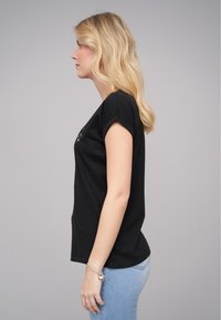 Svart bomull T-skjorte med korte rullede ermer, tilpasset design, detalj i sidesømmen, brukt med lyseblå denimjeans. Jevn tekstur.
