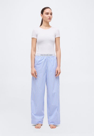 Νεαρή γυναίκα στέκεται ξυπόλυτη φορώντας εφαρμοστή λευκή μπλούζα με κοντά μανίκια και άνετο παντελόνι με κάθετες μπλε και λευκές ρίγες.