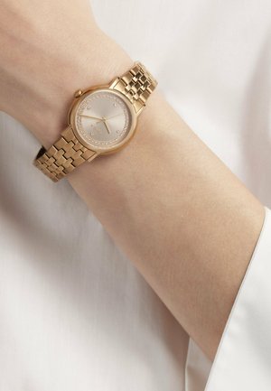 Goldfarbene Armbanduhr mit Kristallakzenten auf einem Gliederarmband aus Metall, getragen am Unterarm vor einem Hintergrund aus einem weißen Langarmhemd.