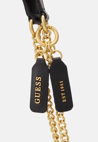 Anse de sac à main en cuir noir avec accents de chaîne dorée et étiquettes gravées avec "GUESS" et "EST 1981." Détails en métal doré mis en valeur.