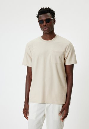Beige T-Shirt mit kurzen Ärmeln, rundem Halsausschnitt und einer Brusttasche. Der Stoff wirkt glatt und leicht und wird mit weißen Hosen kombiniert.