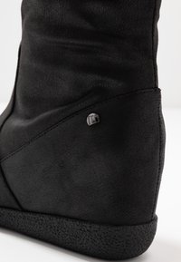 Botte noire montant jusqu'au genou avec tige en cuir souple, détails de design triangulaires et semelle en caoutchouc texturée. Présente un petit logo métallique.