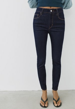 Kvinde iført mørkeblå højtaljede skinny jeans og sorte flipflops, stående mod en ensfarvet lys baggrund.