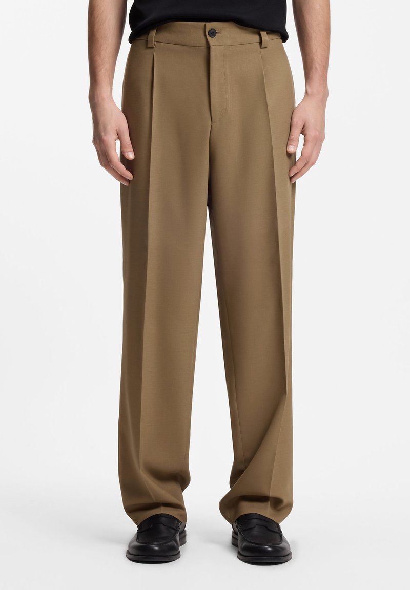 Homme portant un pantalon plissé marron ample avec des mocassins en cuir noir et une chemise noire, debout devant un fond uni.