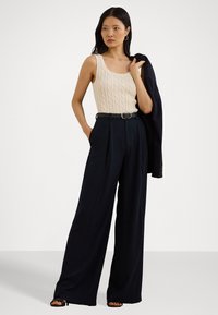 Camisola de malha canelada bege, combinada com calças largas de cintura alta em azul-escuro. Inclui um cinto preto e um casaco azul-escuro pousado sobre um ombro.