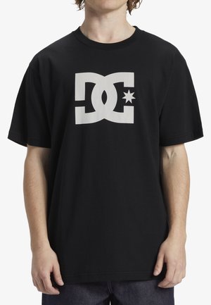 Person trägt ein schwarzes kurzärmeliges Shirt mit einem großen weißen DC-Logo, das in der Mitte auf der Brust einen Stern zeigt.