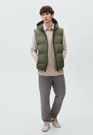 Chaleco de plumas verde oliva con capucha, usado sobre una camiseta polo beige. Jeans grises y zapatillas beige completan el conjunto. Diseño simple, ajuste casual.