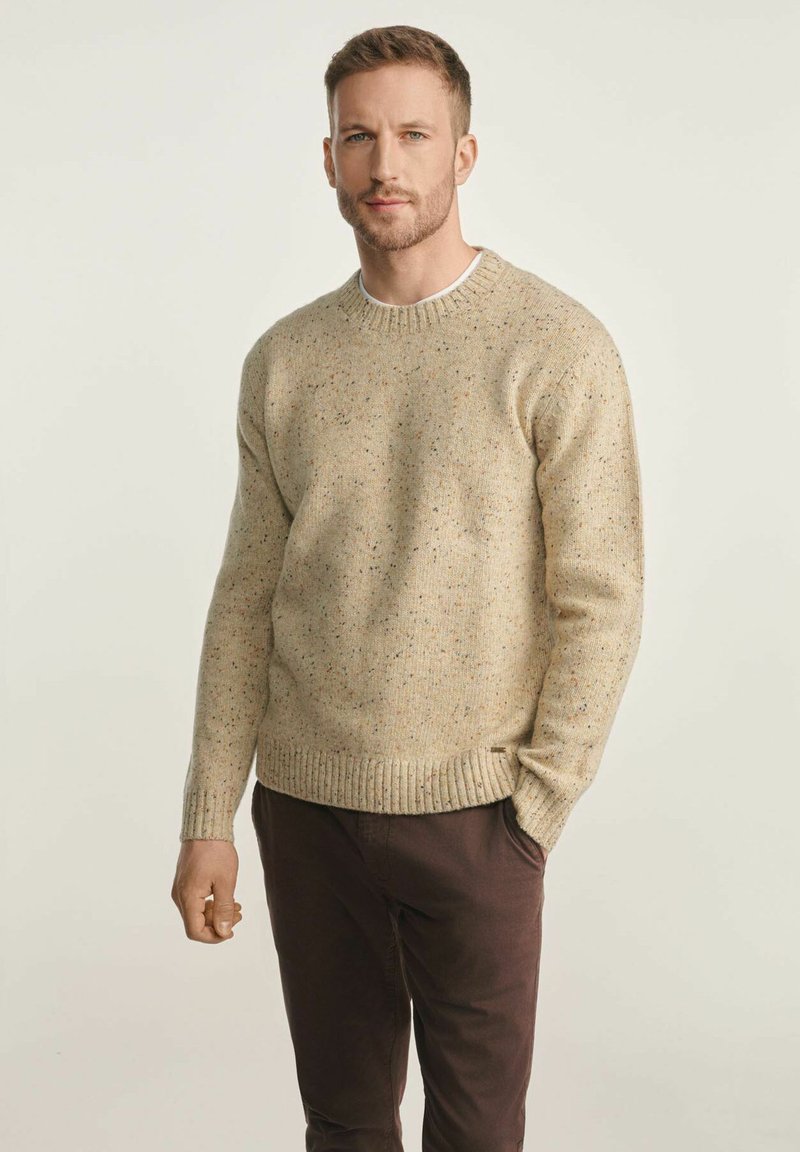 Beżowy sweter dzianinowy o teksturze z drobnymi plamkami, z okrągłym dekoltem oraz ściągaczami przy mankietach i na dolnym brzegu. Zestawiony z ciemnobrązowymi spodniami. Prosty, casualowy design.