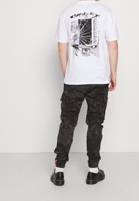 Valkoinen t-paita, jossa musta graafinen kuvio takana, yhdistettynä mustiin maastokuvioisiin cargo-housuihin ja mustiin nauhallisiin kenkiin, neutraalilla taustalla.
