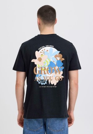 Mann trägt schwarzes T-Shirt mit bunten Blumen und dem Text "Grow as you go" auf der Rückseite, kombiniert mit blauer Jeans, Rücken zur Kamera gewandt.