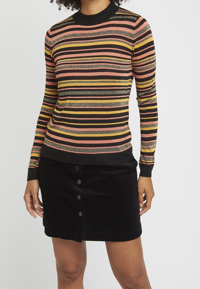 Personne portant un pull à manches longues rayé en noir, rose, jaune et paillettes, assorti d'une jupe en velours côtelé noire à boutons sur le devant.