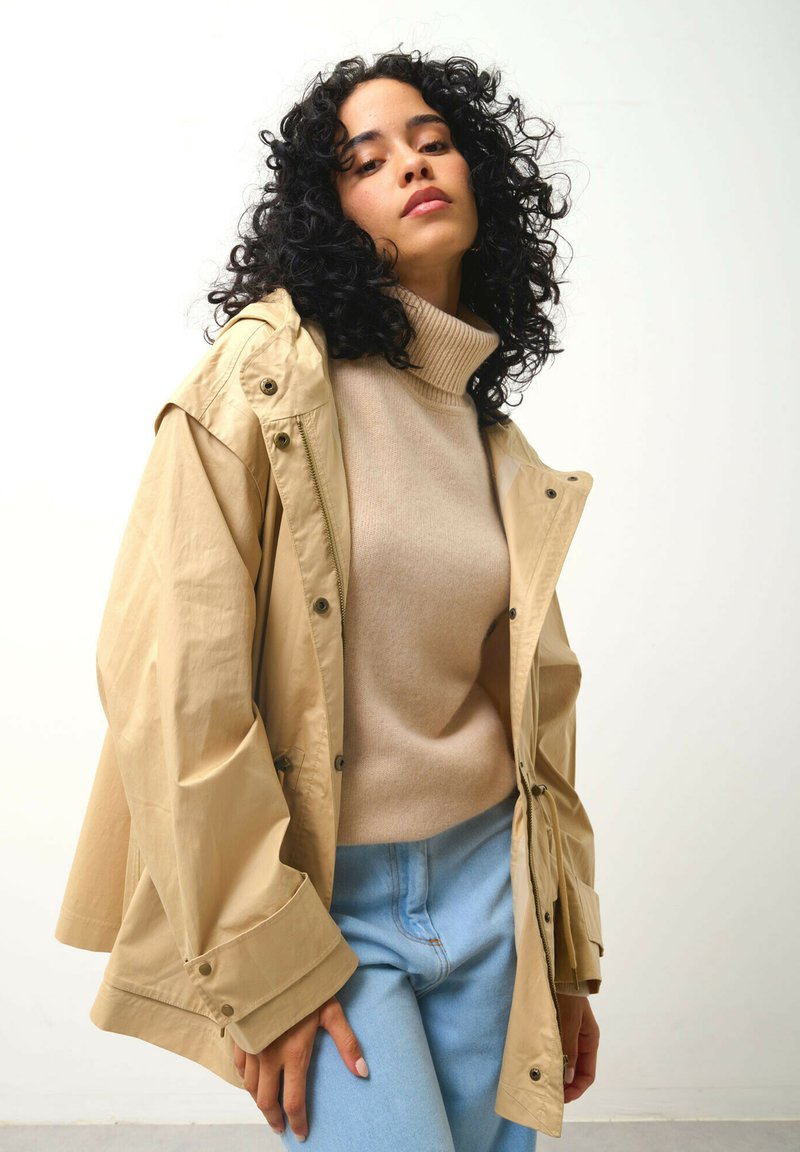 Beige wasserdichte Jacke mit Kapuze über einem hellbraunen Rollkragenpulli. Das Model trägt hellblaue Jeans und posiert mit einer Hand auf ihrer Hüfte.