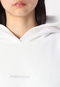 Sweat à capuche blanc avec une poche kangourou, une texture douce et une capuche. Présente un logo "PEGADOR" subtil en gris clair sur le devant.