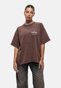 Brązowy, oversize t-shirt z krótkim rękawem, z białym haftowanym logo. Gładka bawełniana faktura, minimalistyczny design i luźny krój.