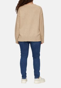 Beige gebreide trui met een loose fit, verlaagde schouders en geribbelde zoom, gecombineerd met blauwe skinny jeans en witte sneakers.