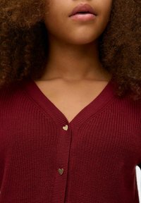 Vyno raudonas megztinis su V formos iškirpte, nugarinė dalis su auksiniais širdelės formos mygtukais priekyje ir tekstūruota medžiaga.