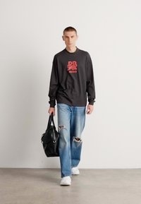 Maglietta nera a maniche lunghe con logo grafico rosso, jeans blu strappati, sneakers bianche e una borsa a spalla nera. Outfit semplice e casual.