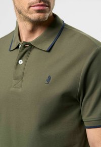 Homme portant un polo vert olive avec une bordure de col marine, deux boutons et un petit logo brodé sur la poitrine.