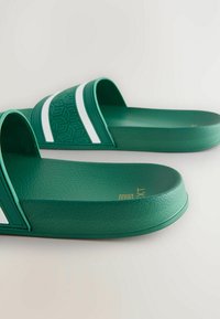 Grüne Rutschsandalen aus strukturiertem Material, mit zwei weißen Streifen auf dem Riemen, abgerundeten Zehen und einem flachen Design.