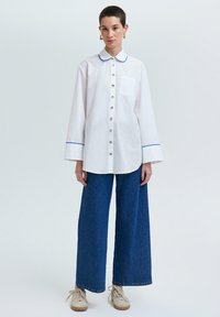 Camisa blanca de botones con cuello y puños azules, combinada con pantalones de mezclilla de pierna ancha. Se aprecian detalles de botones y un bolsillo en el pecho.