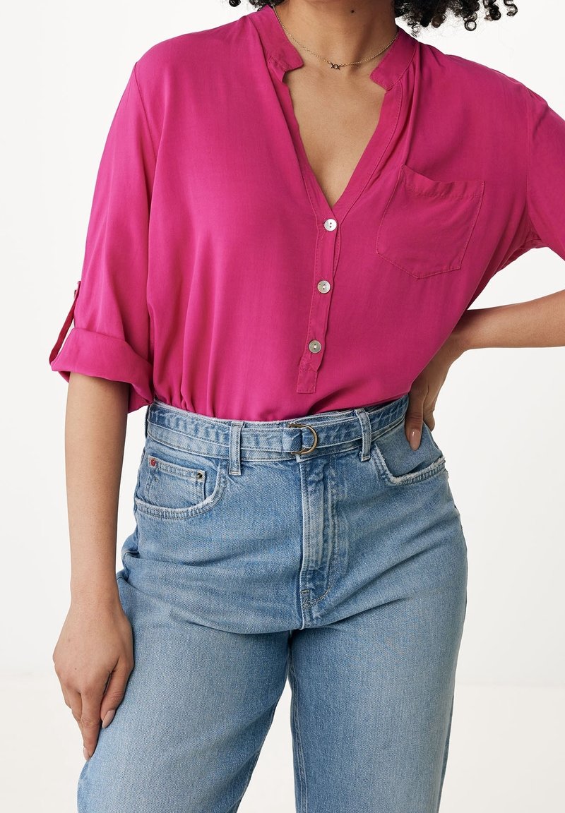 Femme portant un chemisier rose vif à boutons avec manches retroussées, rentré dans un jean taille haute bleu clair avec une ceinture.
