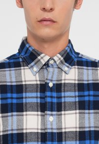 Camisa de flanela com padrão xadrez em azul-marinho, cinza e azul. Colarinho com botão, dois botões na paleta, e textura suave.