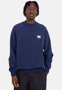 Sudadera azul marino con un cuello y puños acanalados, que presenta un pequeño parche de logo bordado en el área izquierda del pecho. Textura suave.