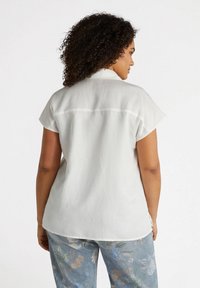 Camicetta bianca a maniche corte con una texture morbida e leggera, caratterizzata da una cucitura a yoke sul retro e un orlo arrotondato, indossata con pantaloni floreali blu a motivi.