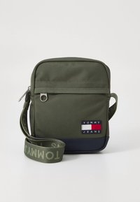 Mala bandoleira de lona verde-azeitona com um bolso frontal com fecho éclair, base azul-marinho e uma alça de marca com o texto "TOMMY". Emblema do logotipo na frente.