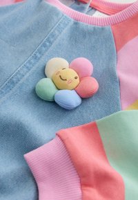 Kleurrijke trui met denim textuur, voorzien van een pastel bloemenapplicatie met een glimlachend gezicht, geaccentueerd door roze, muntgroene en lavendelkleurige boorden.