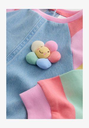 Színes pulóver farmeres textúrával, pasztell virág applikációval, amely mosolygós arcot ábrázol, rózsaszín, menta és levendula színű mandzsettákkal díszítve.