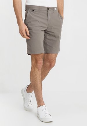 Man wearing gray knee-length shorts and white sneakers walking with hands relaxed, one in pocket, against a plain white background.
