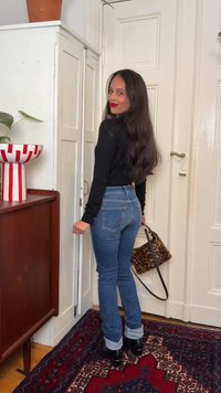 Sieviete ar gariem tumšiem matiem, tērpusies melnā topā, zilās džinsās un melnos apavos, stāv iekštelpās un tur leopardā druku somiņu.