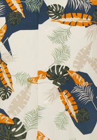 Camisa de manga corta de tela color crema con patrones de hojas tropicales en verde, azul marino y naranja. Tapeta de cinco botones con botones blancos.