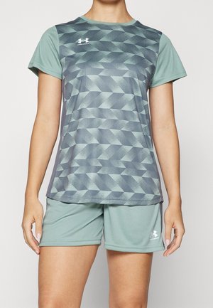 Kobieta w zielonym sportowym t-shircie z geometrycznym wzorem i dopasowanych zielonych szortach sportowych, stojąca z rozluźnionymi ramionami.