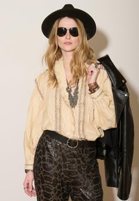 Blouse rayée beige avec un col volants, ornée de broderies sombres. Veste en cuir noire sur le bras. Pantalons imprimé léopard en sequins. Chapeau noir et lunettes de soleil.