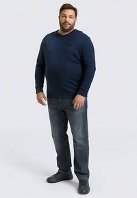 Marineblaues Langarmshirt mit Rundhalsausschnitt, kombiniert mit ausgewaschenen blauen Jeans und dunklen Turnschuhen. Einfaches Design, keine sichtbaren Muster oder Logos.