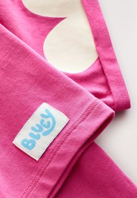 Tessuto di cotone rosa con un grande motivo a cuore color crema, con un'etichetta con il logo "Bluey" blu cucita sull'orlo.