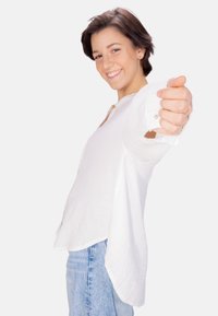 Weißes, strukturiertes Hemd mit abgerundetem Saum und geknöpften Manschetten, kombiniert mit hellblauen Jeans. Das Model posiert mit einem entspannten Ausdruck.