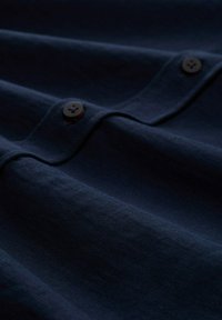 Navyblauwe stoffen shirt met een gladde textuur, voorzien van knopen en zichtbare gestikte details, plat gelegd om plooien en contouren te tonen.